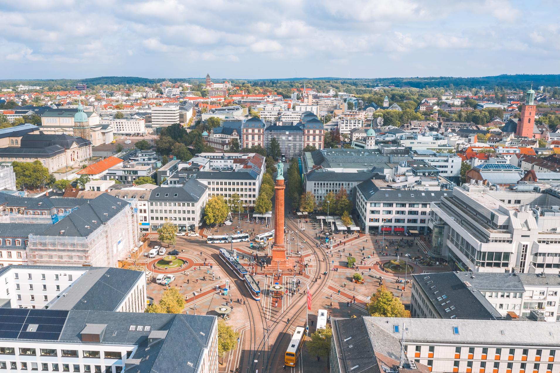 Blick auf eine belebte Innenstadt mit Platz, Straßenbahnen und modernen Gebäuden unter einem bewölkten Himmel.